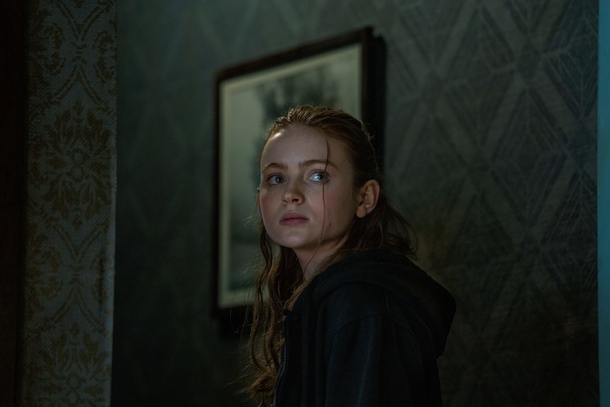 Young white girl with long red hair hair and blue eyes wears a dark hoodie and stands in a gloomy wall-papered hallway.