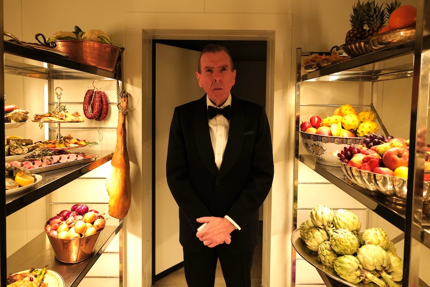 A 60-something man in black tie stands sombrely in a walk-in freezer, the shelves piled with fruit, vegetables and sliced meat