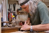A man with long grey hair holds a tool to a piece of timber in a workshop.