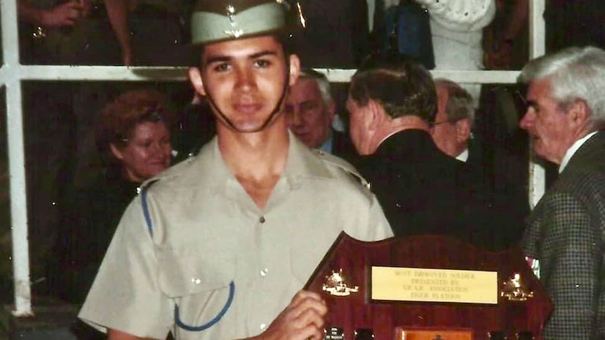 A young man in cadet uniform holding a large wooden plaque with gold metal entries on it.