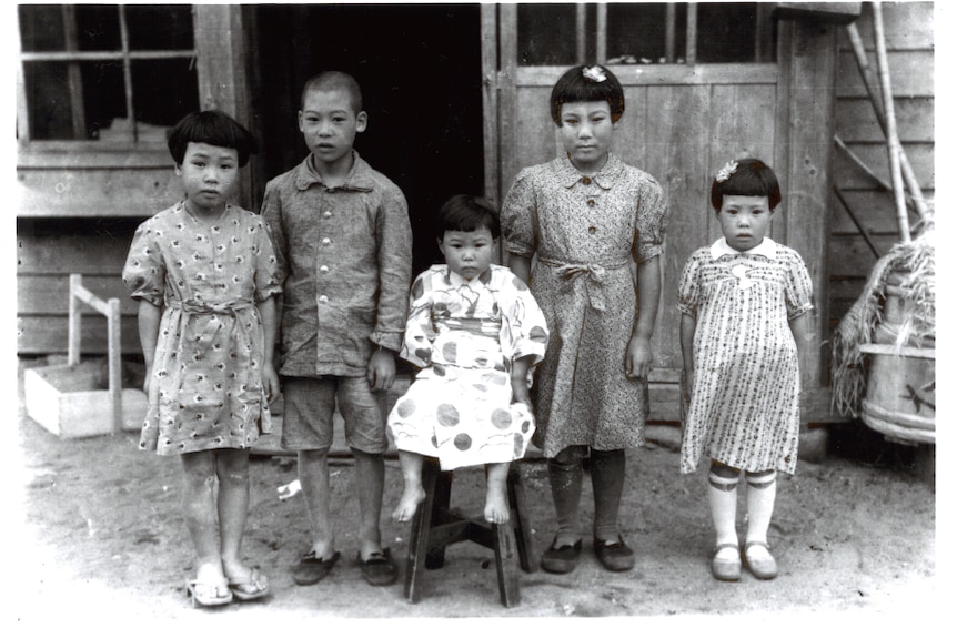 Старая задняя и белая фотография японского мальчика с четырьмя молодыми японскими девочками возле деревянного дома.