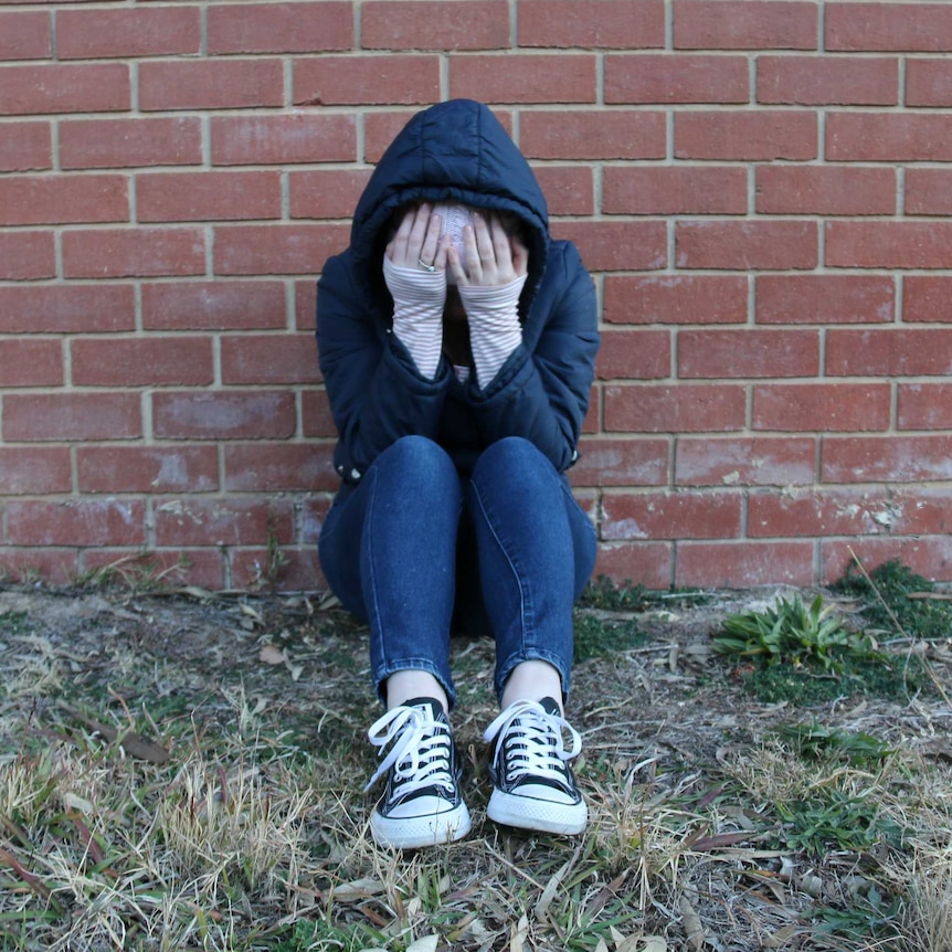 A young person wearing a hood and beanie sits with their head in their hands against a brick wall.