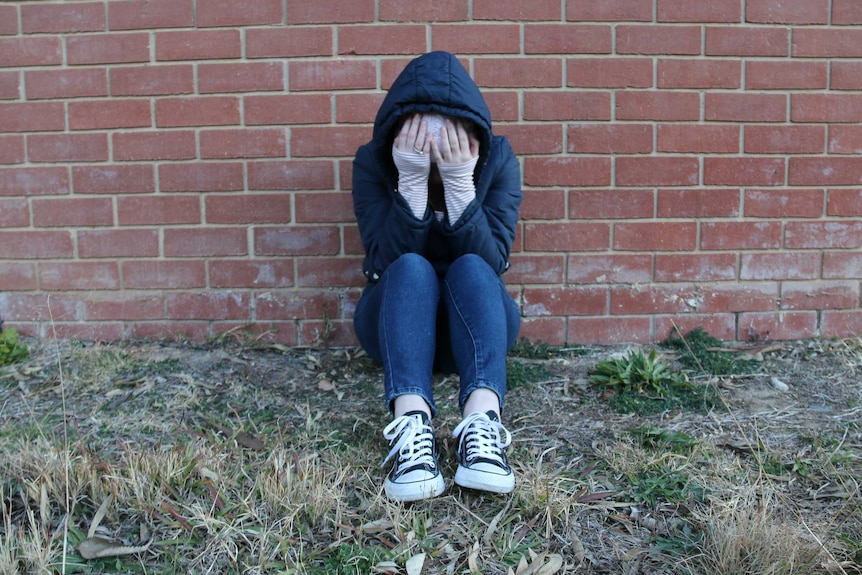 A young person wearing a hood and beanie sits with their head in their hands against a brick wall.