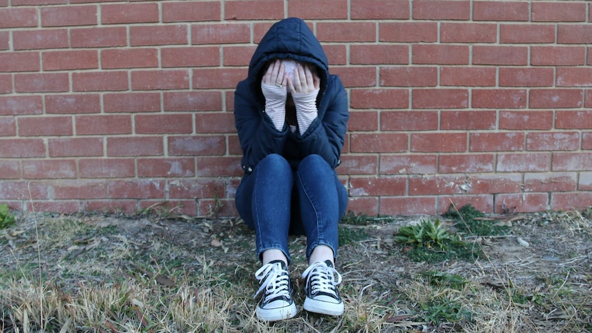 A young person wearing a hood and beanie sits with their head in their hands against a brick wall.