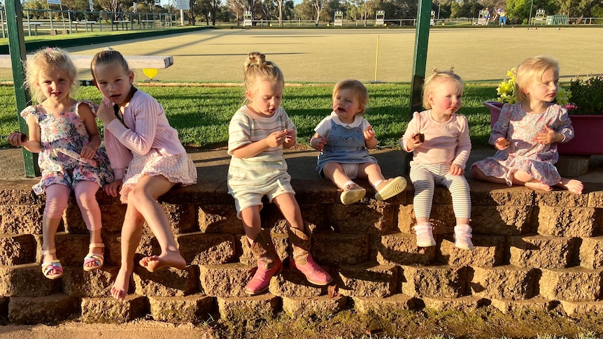 a group of young children sit in a row on a wall, eating.