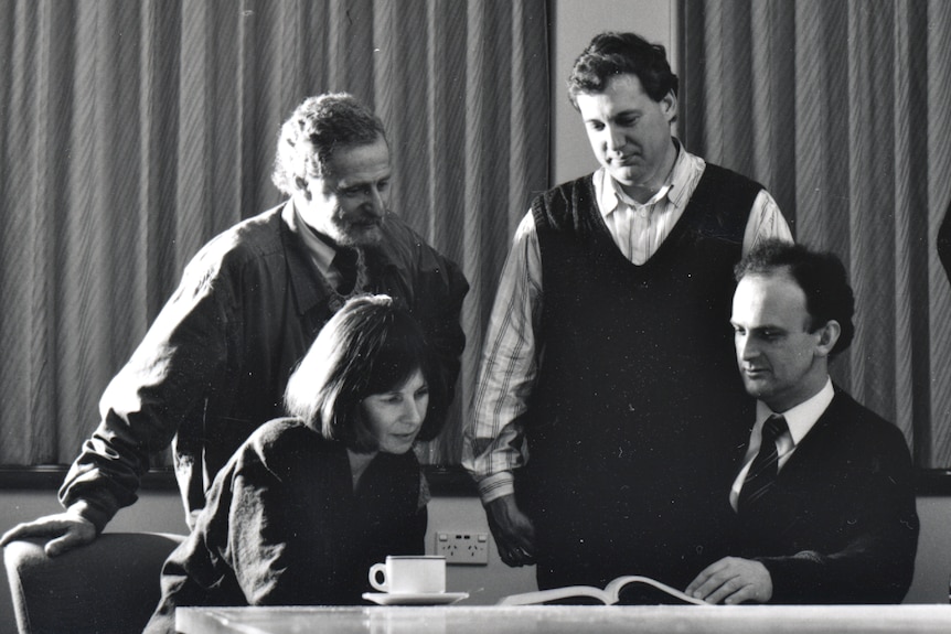 A black-and-white photo of Ian Gust, Suzanne Crowe and other researchers poring over a book on a table.