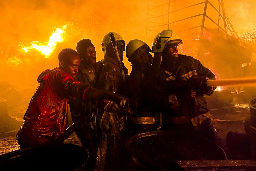 A firefighter holds a hose expelling water, next to other firefighters and volunteers who are all surrounded by flames.