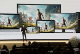 A man speaks on stage in front of a gigantic screen that shows different media devices hosting an Assassins Creed screenshot.