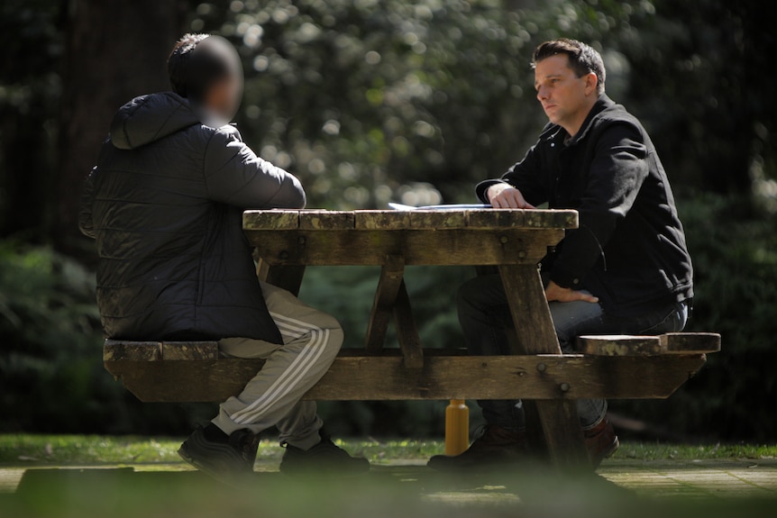 Man wearing jacket and sweatpants sitting at a bench speaking to reporter Michael Atkin.