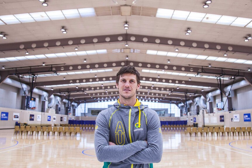 Mitch Mooney stands arms folded on an indoor netball court with a high ceiling.