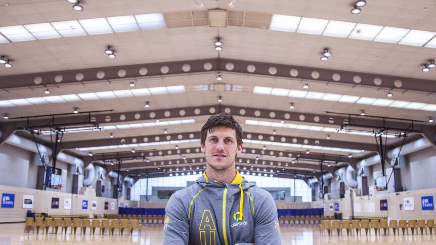 Mitch Mooney stands arms folded on an indoor netball court with a high ceiling.