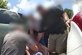 plain-clothes police officers arrest a man between two vehicles. everyone's faces are blurred