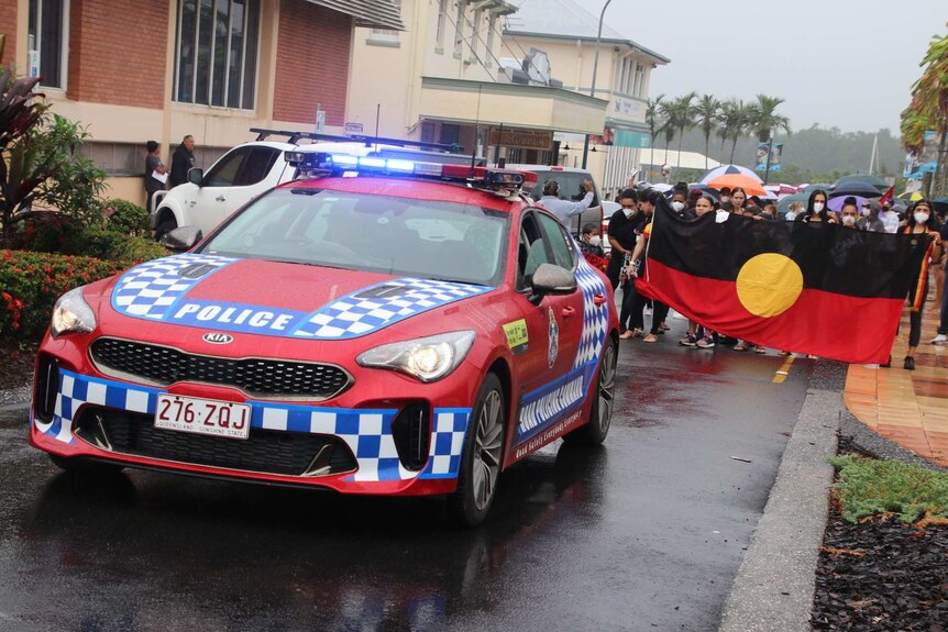 A police car leads a march of protesters who hold up an Aboriginal flag.