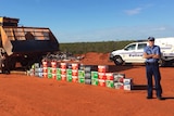 Sergeant Matthew Hartfield stands on red dirt next to around 30 cartons of alcohol as a bulldozer lines up to destroy it.