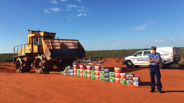 Sergeant Matthew Hartfield stands on red dirt next to around 30 cartons of alcohol as a bulldozer lines up to destroy it.