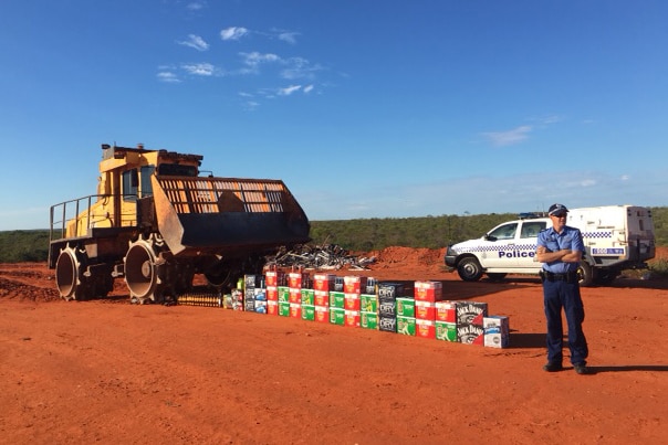 Sergeant Matthew Hartfield stands on red dirt next to around 30 cartons of alcohol as a bulldozer lines up to destroy it.