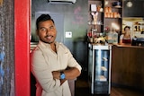 Bhairab leaning against a red pillar on a black wall looking at the camera inside a restaurant.