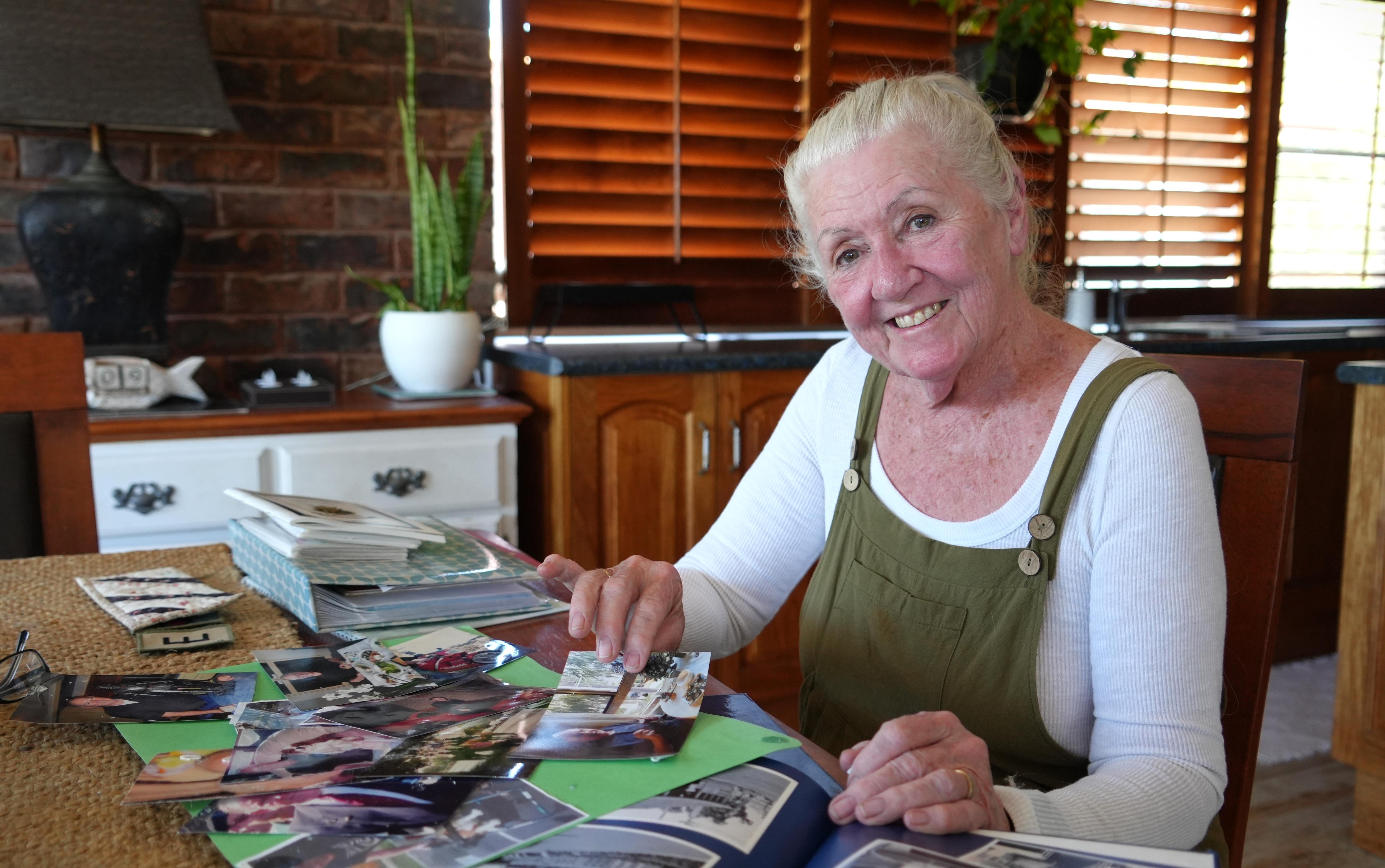 A mature woman wearing overalls sits at a table inside smiling at the camera, holding photos and clippings.