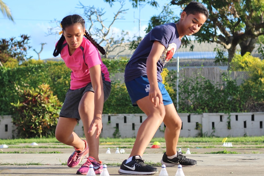 A teenage boy and girl run side by side toward camera, about to crouch and pick up a shuttlecock