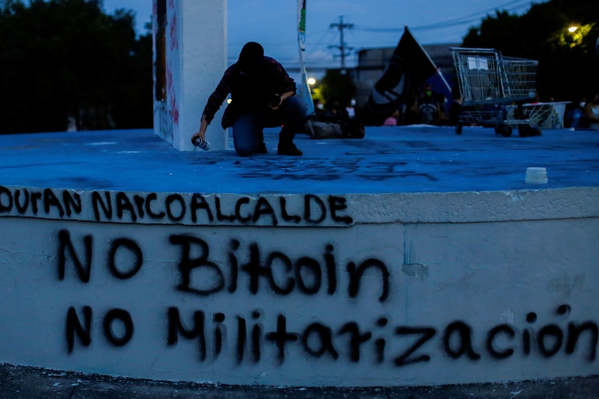  A man dressed in black takes part in a protest, spray-painting no bitcoin on a public wall at night.