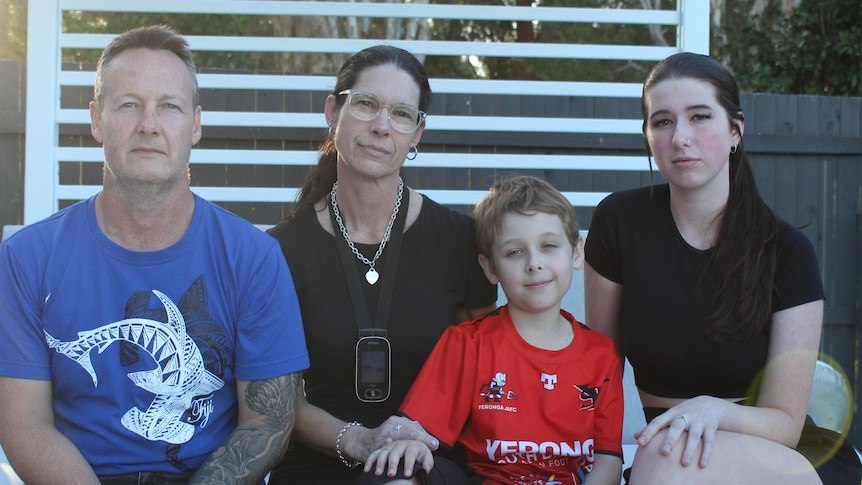 An image of Heath family sitting on pool lounge with white pergola and brown fence in background 