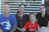 An image of Heath family sitting on pool lounge with white pergola and brown fence in background 