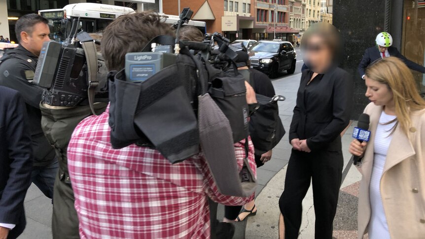 A woman, face blurred, is surrounded by the media outside court.