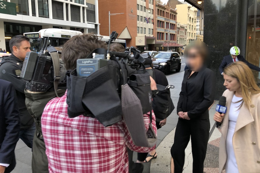 A woman, face blurred, is surrounded by the media outside court.