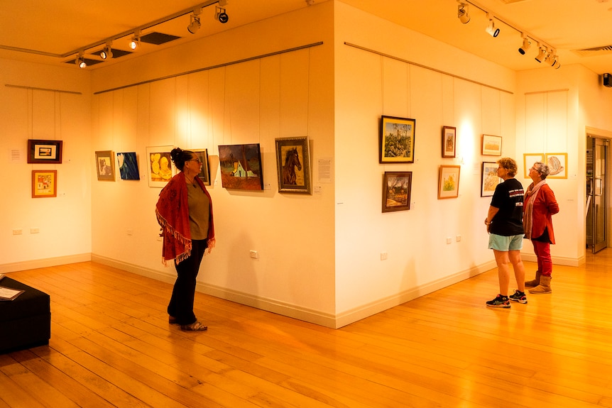 Three people stand in and art gallery setting looking at work.