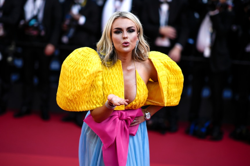 Tallia Storm blowing a kiss wearing a dress with big puffy yellow sleeves, a pink bow and a pale blue skirt. 