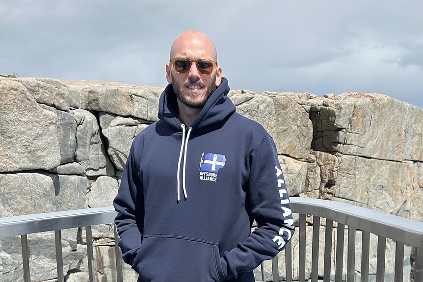man in navy hood jumper and sunglasses stands in front of a rock wall 