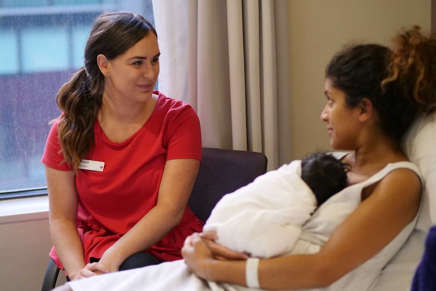 A midwife sits in a red uniform and chats to a woman on a hospital bed with a baby sleeping on her chest.