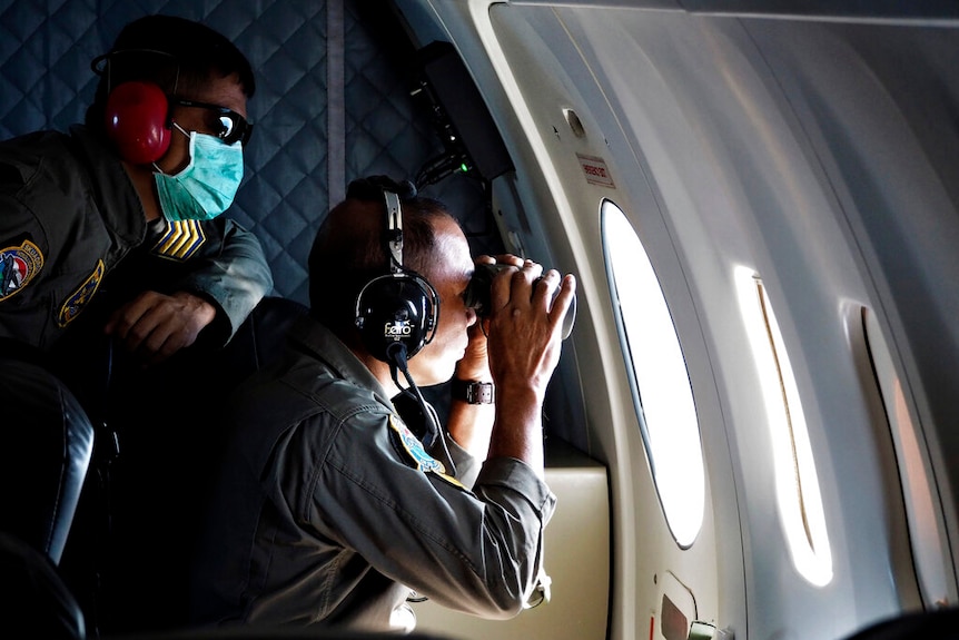Two soldiers in an airplane look out of a circulate window, with one looking through binoculars.