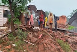 People with umbrellas stand amidst the rubble of a brick building. 