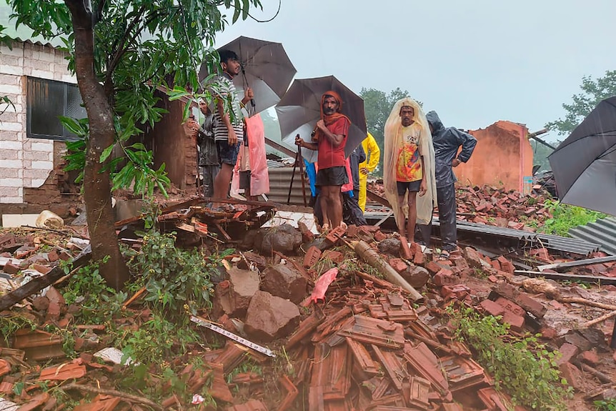 People with umbrellas stand amidst the rubble of a brick building. 