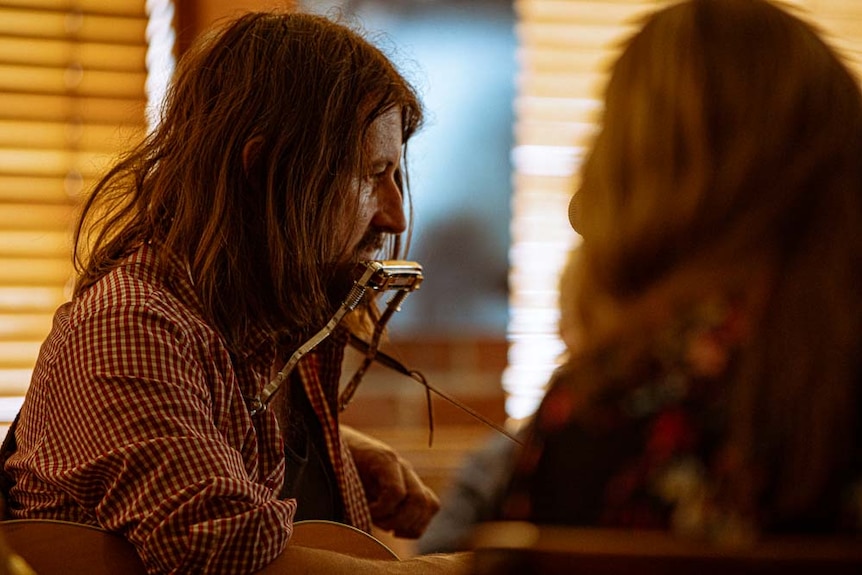 A man with long hair sits playing guitar and harmonica