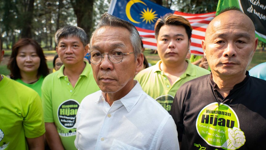 A man in a business shirt and glasses is surrounded by people in green-t shirts with a Malaysian flag in the background