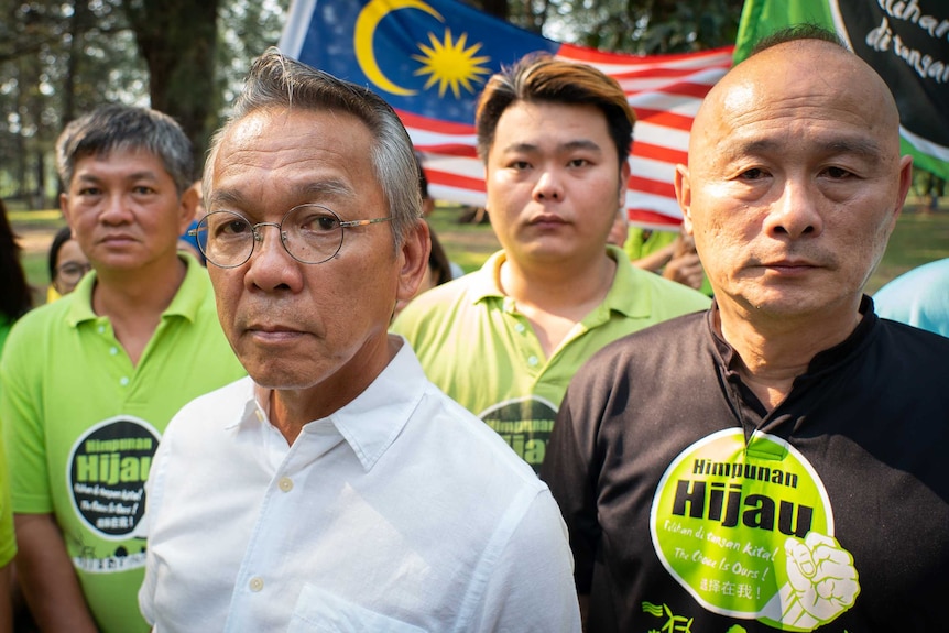 A man in a business shirt and glasses is surrounded by people in green-t shirts with a Malaysian flag in the background