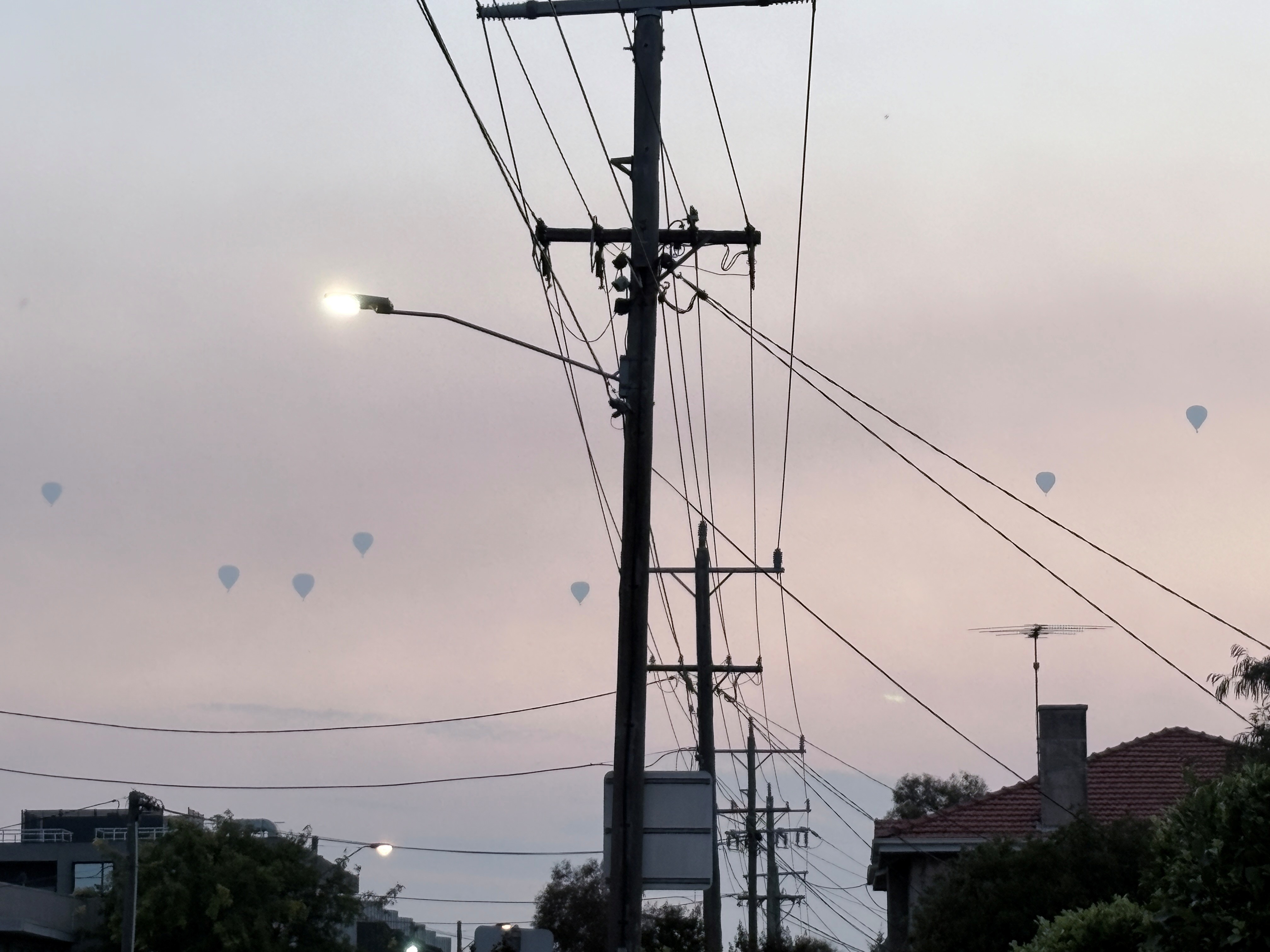 Sete balões de ar quente aparecem como silhuetas cinza-claras contra um céu cheio de fumaça marrom e creme sobre casas e linhas de energia.
