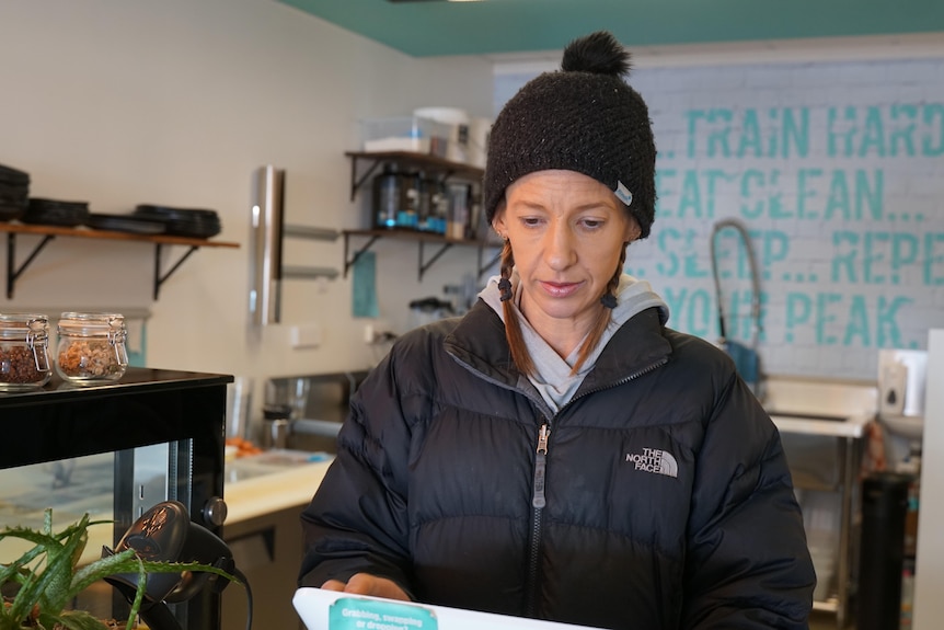 A woman in a black beanie and puffer jacket works a till in a cafe.