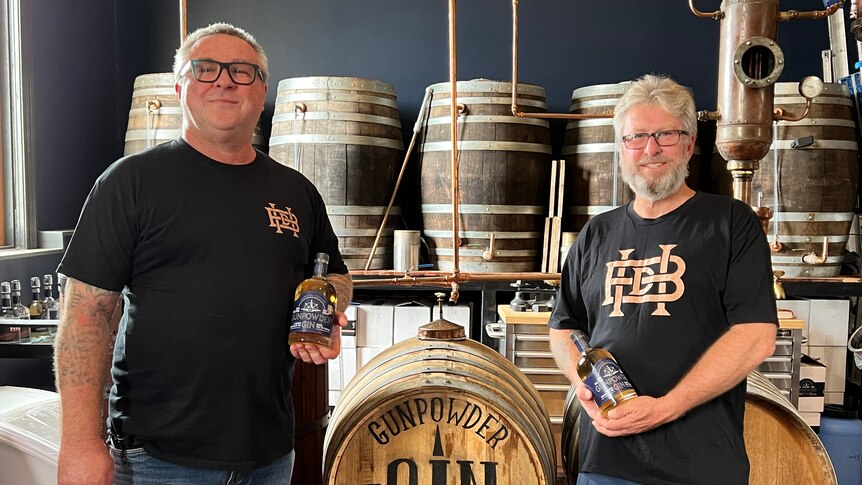 Two men stand in front of a barrel holding bottles of gin.