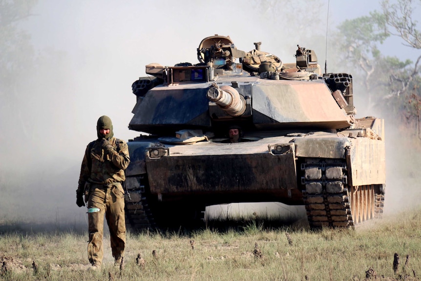An Australian soldier walks in front of a M1A1 Abrams main battle tank in scrubland.