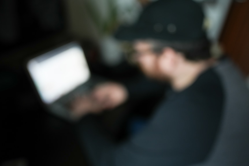 A man sits in front of a laptop he is out of focus and you cannot make out his feature