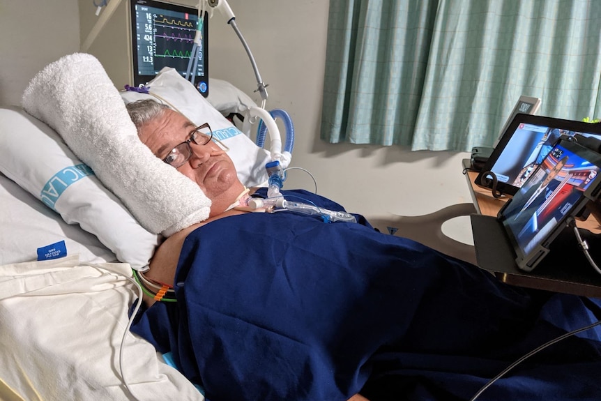 A man with breathing support lies in a bed in hospital watching screens