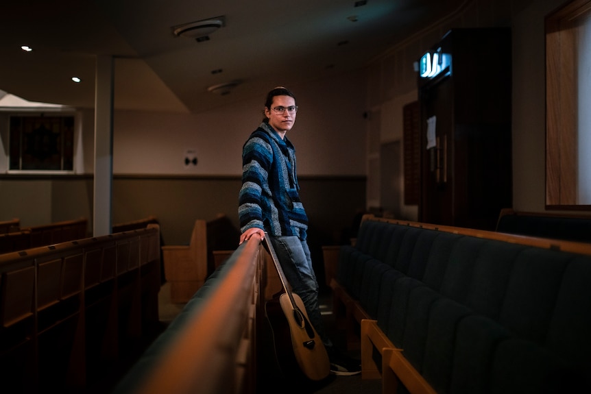 Avishai Conyer stands inside a synagogue, leaning on a bench with his guitar.