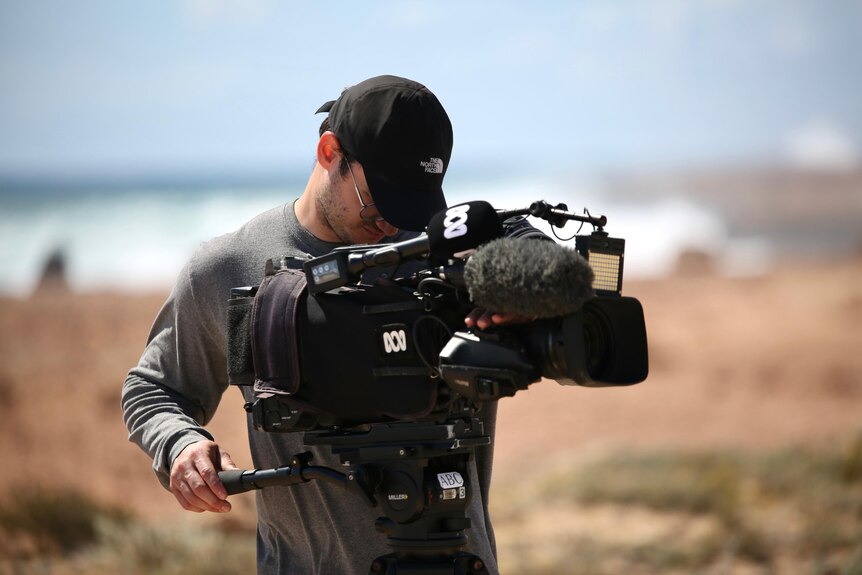 A man wearing a hat and grey t-shirt operates an ABC branded video camera on a rugged cliff near a wild ocean.