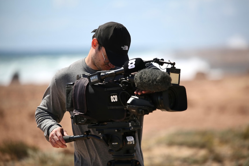 A man wearing a hat and grey t-shirt operates an ABC branded video camera on a rugged cliff near a wild ocean.