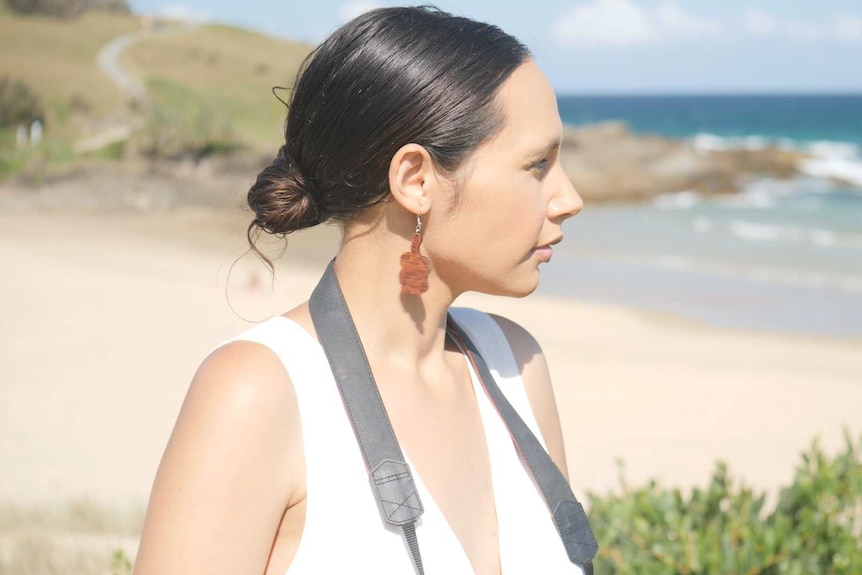 A young woman at the beach looking at the ocean with a camera around her neck.