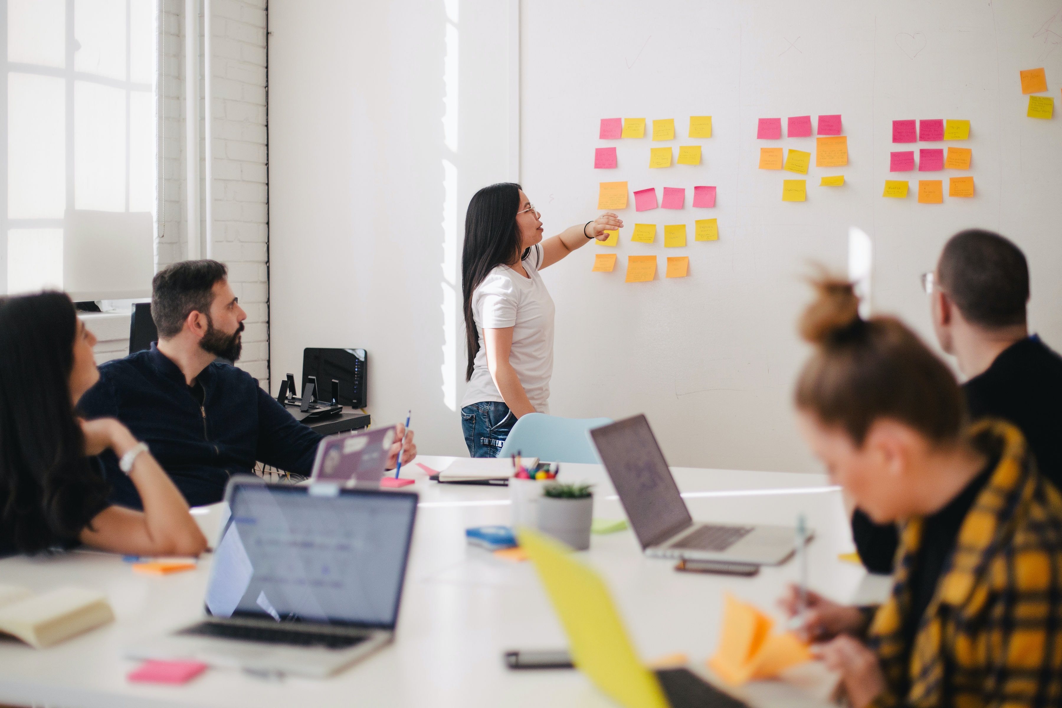 A short young woman of Asian appearance hosts a meeting with sticky notes on a wall. People around a table take notes around her