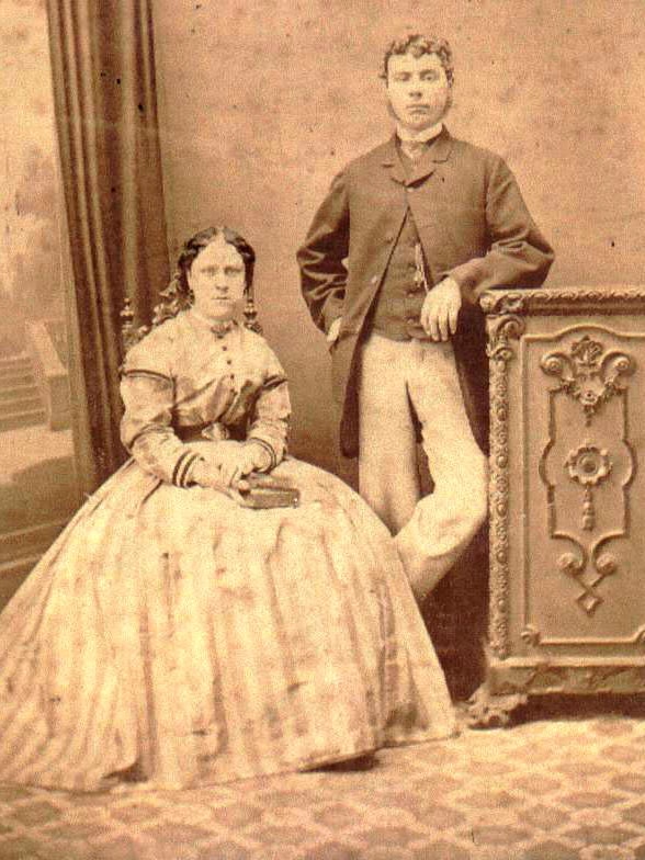 A black and white photo of a man standing next to a seated woman, both in Victorian garb 
